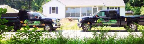 Two black Kent Tree branded pickup trucks parked side by side on a residential street in front of a white house, small dirt pile in the yard.